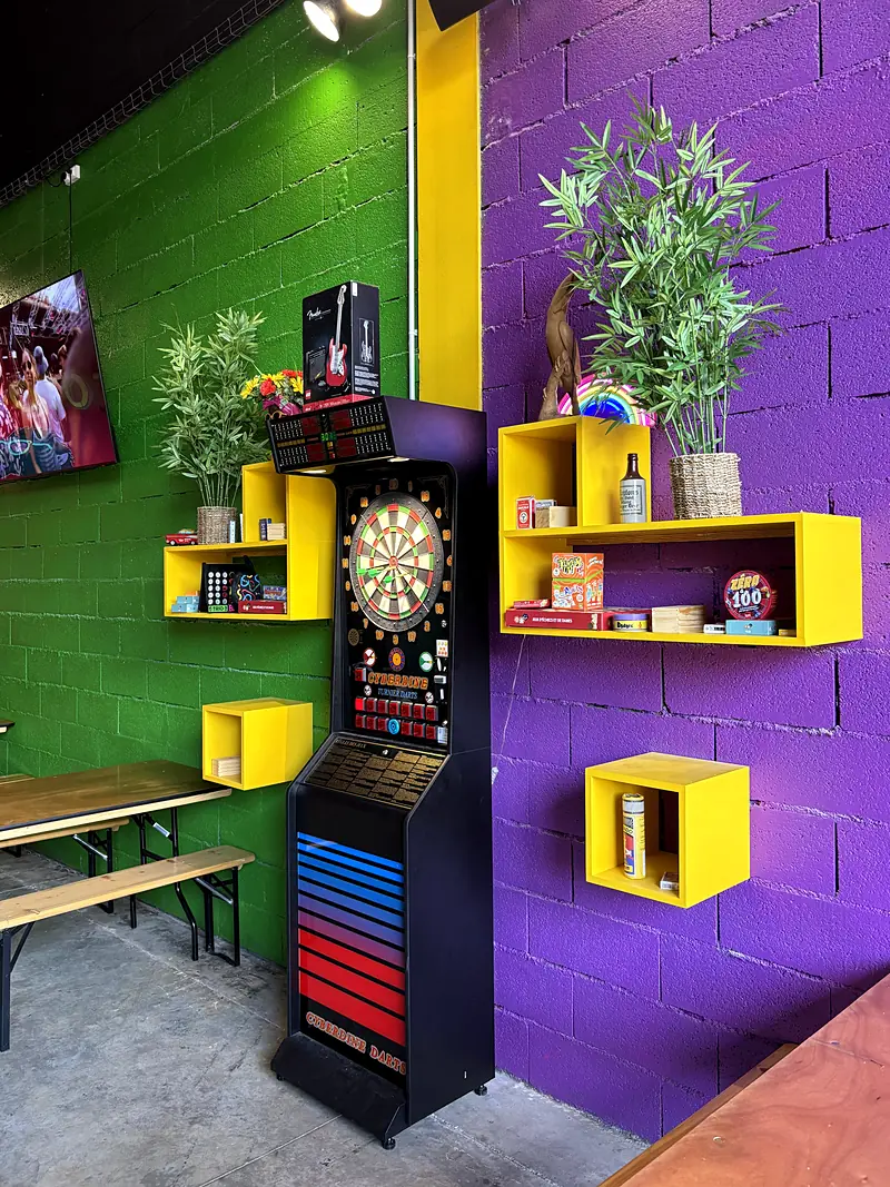 La machine de fléchettes devant les murs vert et violet, avec les étagères jaunes.